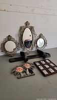 Group shot of three ornate vintage mirrors, two black wall shelves, two palm tree picture frames, and a wooden multi-photo frame placed on flat surface.