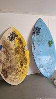 Two ZAP skimboards leaning against a wall, one yellow and white covered in dirt and paint wear, the other light blue with paint splatter and green frog decal.