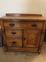 Front view of antique light brown wooden cabinet showing metal drawer handles, paneled door, and wooden caster wheels.
