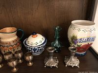 Shelf showing ceramic creamer, sugar bowl, biscotti urn, miniature chalices and metal candlesticks, and green glass vase.