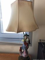 Full view of the Chinese Famille Rose porcelain Magu figurine lamp showing the beige lamp shade and figurine base on a wooden stand.