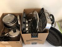 Photo showing multiple metal cookware and bakeware items including pots with lids stacked in a cardboard box, muffin tray, roasting pan with rack, and additional lids