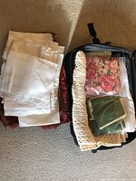 Lot of assorted folded fabrics including white lace-edged napkins, floral prints, crocheted afghan, and other textiles packed partially in a suitcase.
