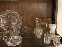 Various clear glass dishes and platters including a cruet, small bowls, candleholders and a ribbed vase arranged on a wooden shelf.