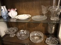 Two shelves with various glassware and ceramic items including vases, bowls, cake plate and basket