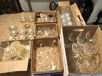 Several boxes containing assorted sets of wine glasses, brandy snifters, martini glasses, rocks glasses, and shot glasses on a table