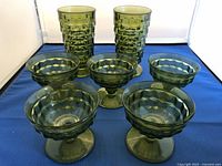 Photo showing 2 tall green textured glasses and 5 green textured footed bowls arranged on a blue surface.