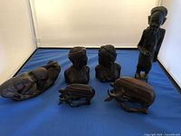 Front view of 6 hand-carved wooden cultural sculptures including human figures and animal forms on a blue cloth background within a lightbox.