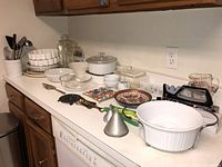 Full view of kitchenware on countertop showing variety of items including Corning Ware, pie plates, bowls, canisters, cutlery, trivets, and sharpener.