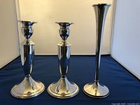 Three sterling silver candle holders shown together against neutral backdrop on blue surface, two shorter identical and one taller slender holder
