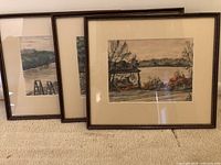Three framed watercolor paintings leaning against a wall on carpet, showing river and dock scenes with wooden frames and matting.