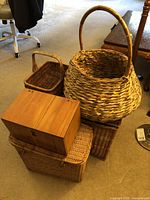 Photo showing five wicker baskets of varied sizes and one wooden box stacked and displayed on carpet indoors. The baskets show traditional weaving with natural wicker material. The wooden box is rectangular and has a simple natural wood finish with a lock.