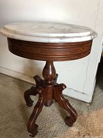 Full view of Victorian style round side table with white marble top and wooden ornate legs against wall.