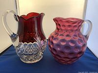 Photo showing two ruby glass water pitchers side by side on white background. The left pitcher is tall with clear handle and faceted base, the right pitcher is round with hobnail texture and clear handle.