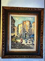 Front view of framed oil painting depicting a street scene with people walking near an archway building.