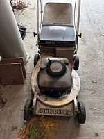 Front view of the John Deere 14PZ lawn mower showing the metal deck, pull cord starter, engine cover, and handle with attached grass catcher bag, highlighting dirt and wear signs.
