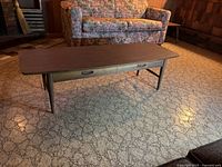 Coffee table positioned in a carpeted living room near floral couch showing side view of laminate top and legs.