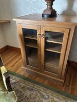 Front angled view of a wood cabinet with glass paneled double doors and brass pulls, showing the three shelves inside.