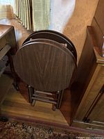 Four stacked round wooden tray tables in brown finish with visible wood grain, shown folded and stored vertically.