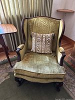 Front view of vintage green wingback chair showing green upholstery, carved wooden legs and frame, and included decorative pillow with floral stripes.