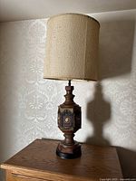 Full view of the vintage table lamp showing the decorative wooden base and beige fabric lampshade on a wooden surface against wallpaper.