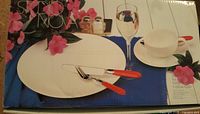 White dish set displayed with plates, tea cup, tea saucer, and cutlery on a blue placemat with pink flowers.