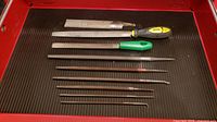 Seven assorted metal files laid on black ribbed tray. Includes two half round files with handles, rat tail files, square file, and two chain saw files.