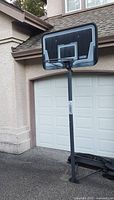 Photo of freestanding adjustable basketball hoop set up outdoors in front of garage, showing backboard, pole, and base.