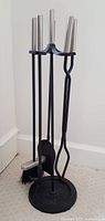 Fireplace tool set with four metal tools on a round stand, silver handles on tools, black metal body and base.