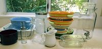 Display of all items on counter under window, showing ceramic colanders stacked, glass beverage dispenser and various jars and baking dishes