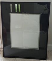 Front view of large black picture frame with glass covering and white matting inside, showing reflective surface and surrounding area reflected in glass.