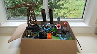 Box with Mega Bloks pieces including brown crane, green baseplates, black funnels, grey barrels, red and white round parts, visible from above near a window.