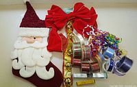 Photo showing a Santa Claus Christmas stocking, large red bow with jingle bells, multiple rolls of specialty tape and ribbons with various patterns and colors including plaid and metallic finishes, curly multi-colored ribbons.