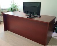 Photo showing full length of solid dark brown office desk with flat top surface and back panel, desk positioned with plant and window in background