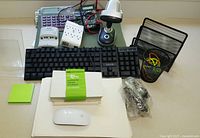 Photo showing various office supplies including a black wired keyboard, white wireless mouse, desktop calculator, black metal mesh document sorter, pen holder with rubber bands, green sticky notes, bundles of white envelopes, assorted cables, white electrical adapter plug, and white power strip.