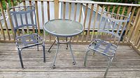Full view of the round glass-topped metal patio table and two matching metal chairs on a wooden deck.