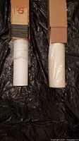 Two rolls of seamless photography backdrop paper in cardboard boxes, one brilliant white and one off white, laid on black plastic sheet.