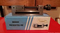 Photo shows the precision tool vise resting on its original blue box labeled 'QGG63 PRECISION TOOL VISE'. The vise is made of metal with a screw mechanism and is in good used condition.