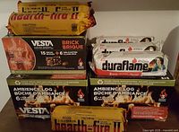 Photo showing the lot consisting of 2 yellow Hearth-Fire II fire log boxes, 6 loose Duraflame fire logs in white packaging, 2 black Vesta fire starter brick boxes, and 2 black Ambience Log fire log boxes.