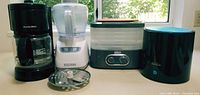 Front view of the four small appliances on a countertop by a window: coffee maker, food processor, food dehydrator, and air humidifier.