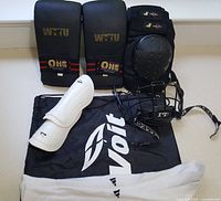 Two pairs of black sparring gloves, white shin guards, black face mask, white socks, and black bag with Volt logo.