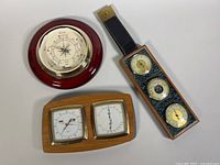 Photo of three vintage barometers/weather station laid on white surface: Fischer weather station with three dials, small wooden mounted twin dial instrument, and a round Thermor barometer.