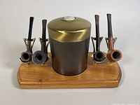 Front view of four tobacco pipes on a walnut display stand with a brown humidor jar with brass lid in the center