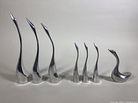Seven polished aluminum swan sculptures arranged in a row, showing varying heights and sleek elongated necks.