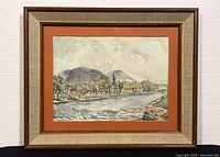 Framed watercolour landscape painting showing a river, houses, trees, mountains, and a church steeple under a cloudy sky with a beige frame and orange matting.
