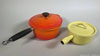 Side view of orange-red Le Creuset cast iron enamel pot with black wood handle beside yellow enamel pot with lid and handle