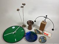 Photo showing multiple items including green BAUXX wall clock, blue ceramic ashtray, metal floral sculpture, wood/metal vase, brass candleholders, and ashtrays