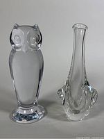Front view of clear crystal owl figurine and elongated crystal vase side by side on a white background.