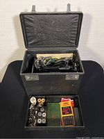 Interior of black carrying case with removable tray showing assorted bobbins, presser feet, Singer needle pack, green Singer booklet and cords