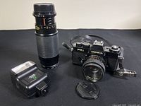 Yashica FR1 camera body with attached 50mm f/1.9 lens, 100-300mm zoom lens standing upright, Sunpak Auto 170 flash and lens cap on black cloth
